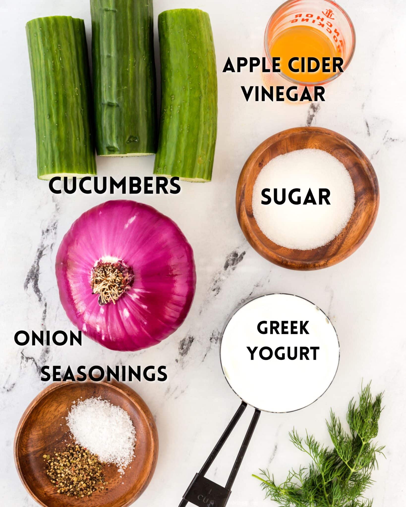 Ingredients for a recipe displayed on a white surface: sliced cucumbers, a halved red onion, a small bowl of apple cider vinegar, a mound of sugar, a bowl of Greek yogurt, salt with seasoning, and fresh dill.