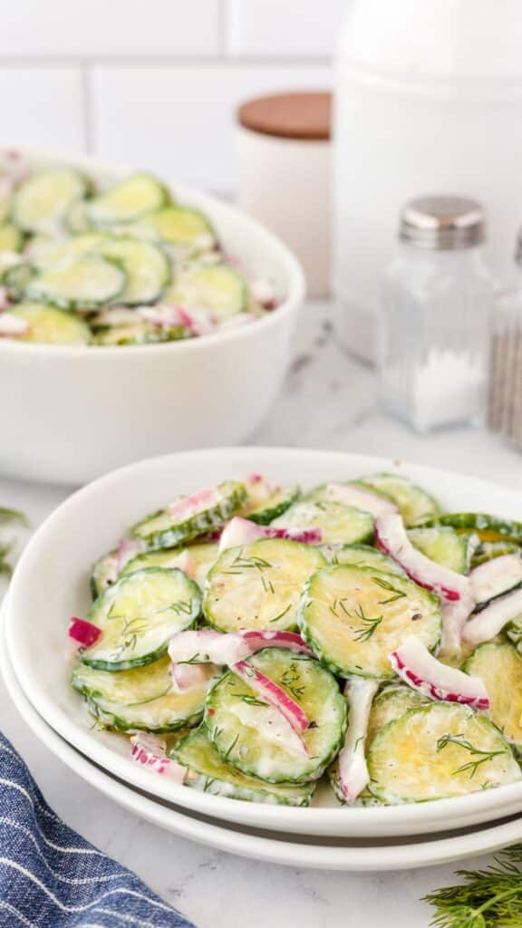 A close-up of a creamy cucumber salad with sliced cucumbers, red onions, and dill in a white bowl. Another bowl of the same salad is blurred in the background. Salt and pepper shakers are visible nearby.