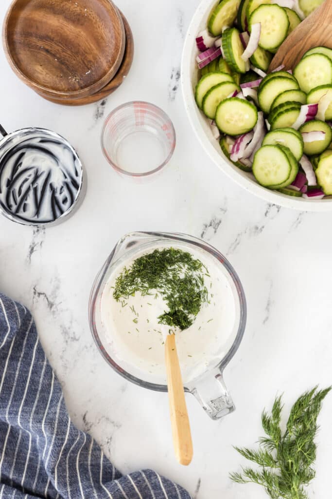 A top-down view of a kitchen counter with bowl of sliced cucumbers and red onions, a measuring cup with yogurt and herbs, a wooden spoon, a wooden dish, and a blue striped cloth. Fresh dill is scattered on the counter.