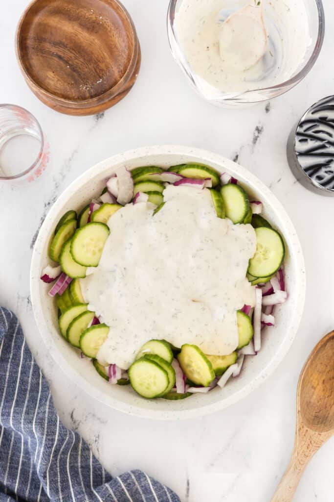 A bowl of cucumber salad with sliced cucumbers, red onions, and creamy dressing. Surrounding the bowl are an empty wooden bowl, a striped towel, a wooden spoon, and a glass bowl with dressing remnants on a marble countertop.