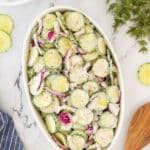 A white oval dish filled with creamy cucumber salad, featuring sliced cucumbers, red onions, and a dill dressing, is placed on a marble surface. Nearby are forks, a salt and pepper set, fresh dill, and a wooden spoon.