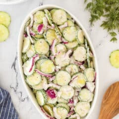 A white oval dish filled with creamy cucumber salad, featuring sliced cucumbers, red onions, and a dill dressing, is placed on a marble surface. Nearby are forks, a salt and pepper set, fresh dill, and a wooden spoon.