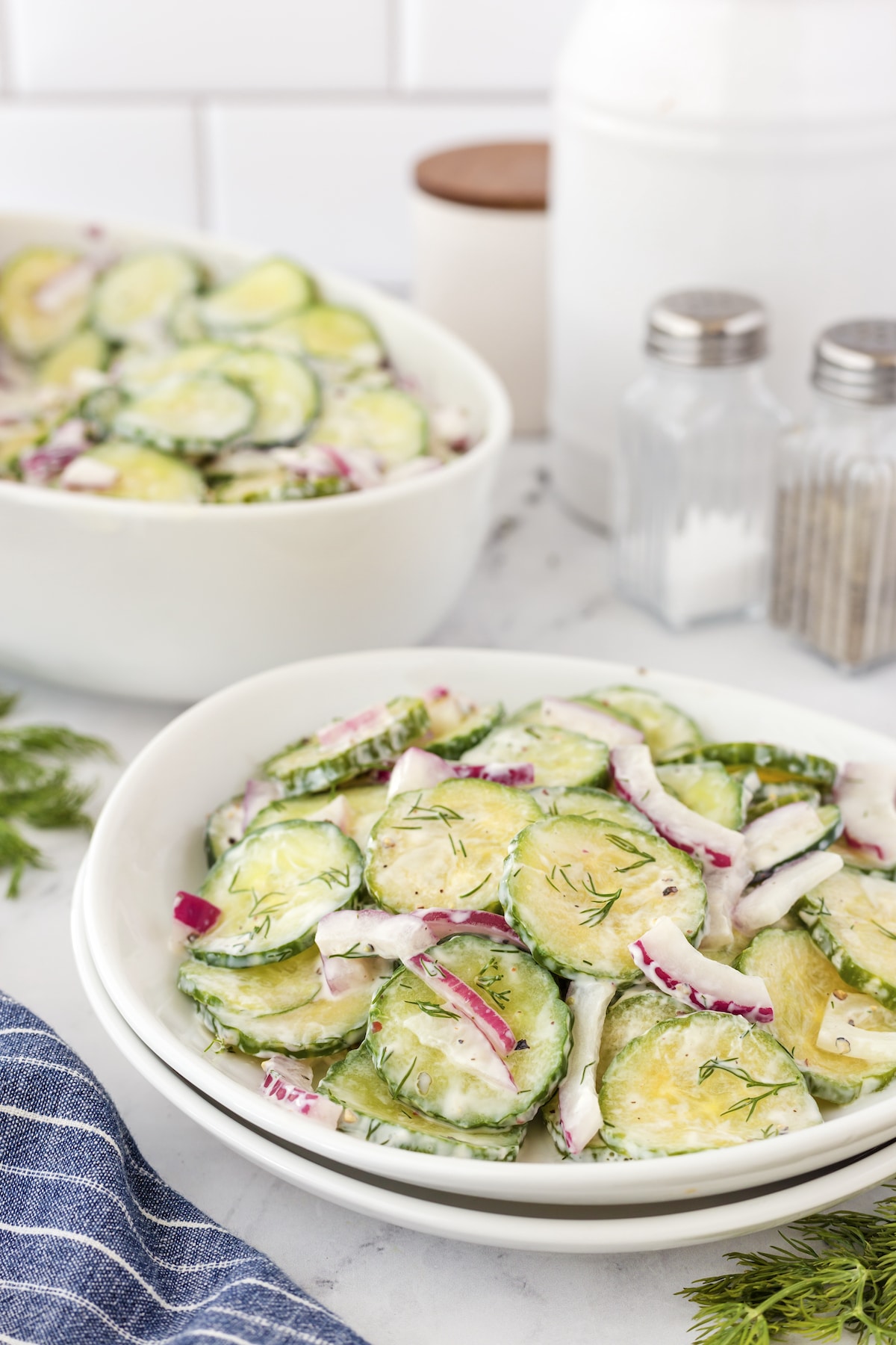 A bowl of creamy cucumber salad with sliced cucumbers, red onions, and fresh dill on a table. A larger bowl with the same salad is in the background along with a salt shaker, a pepper shaker, and a folded blue cloth.