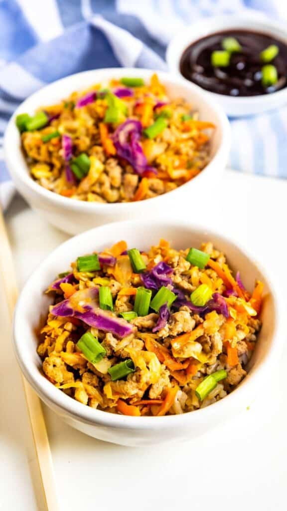 Two white bowls filled with a colorful stir-fry featuring rice, scrambled eggs, chopped carrots, purple cabbage, and green onions. A small dish of dark sauce and a blue-striped cloth napkin are in the background.