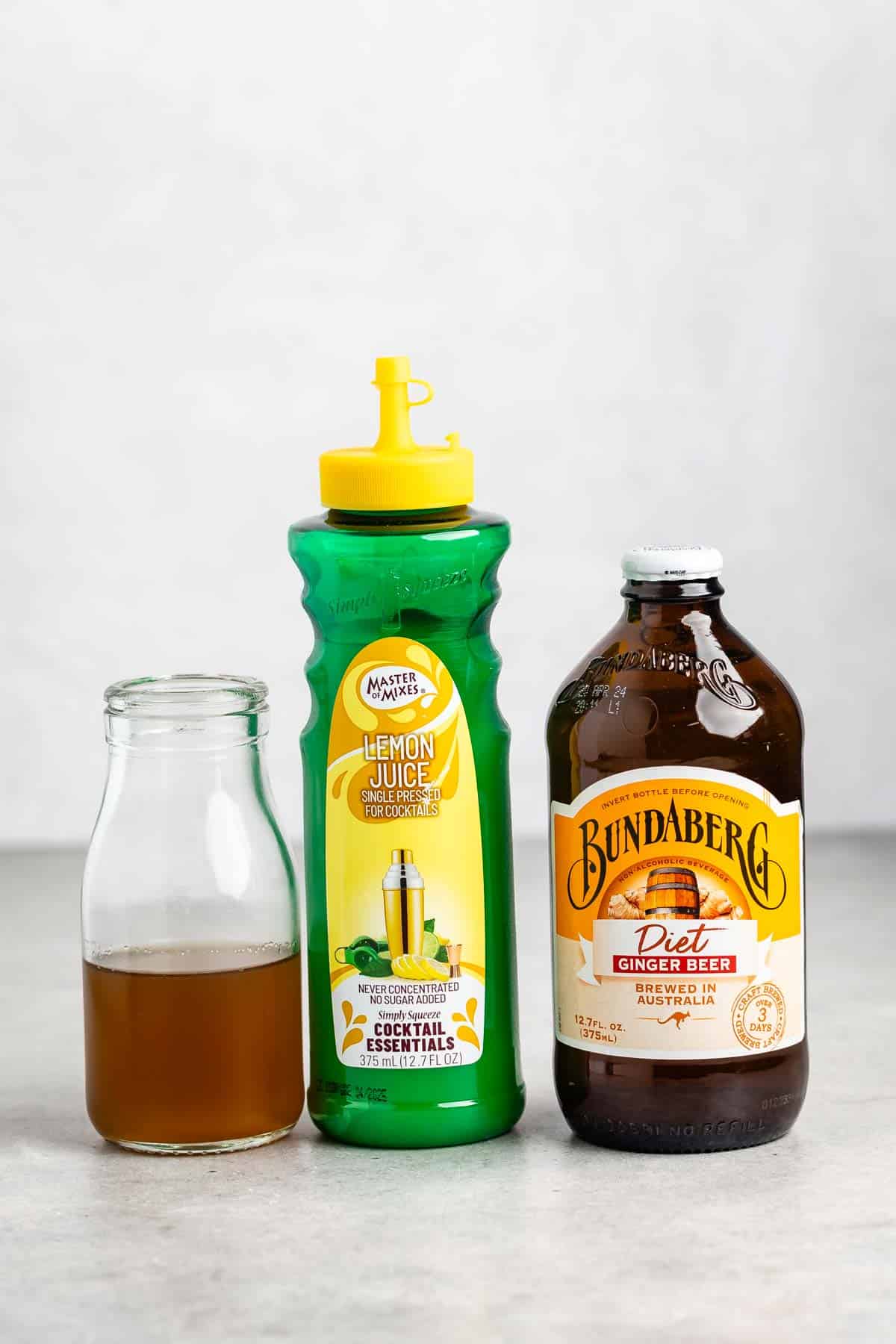 Three bottles are on a counter: a clear glass bottle with brown liquid, a green plastic lemon juice bottle with a yellow cap, and a brown glass bottle of Bundaberg Diet Ginger Beer with a yellow cap.