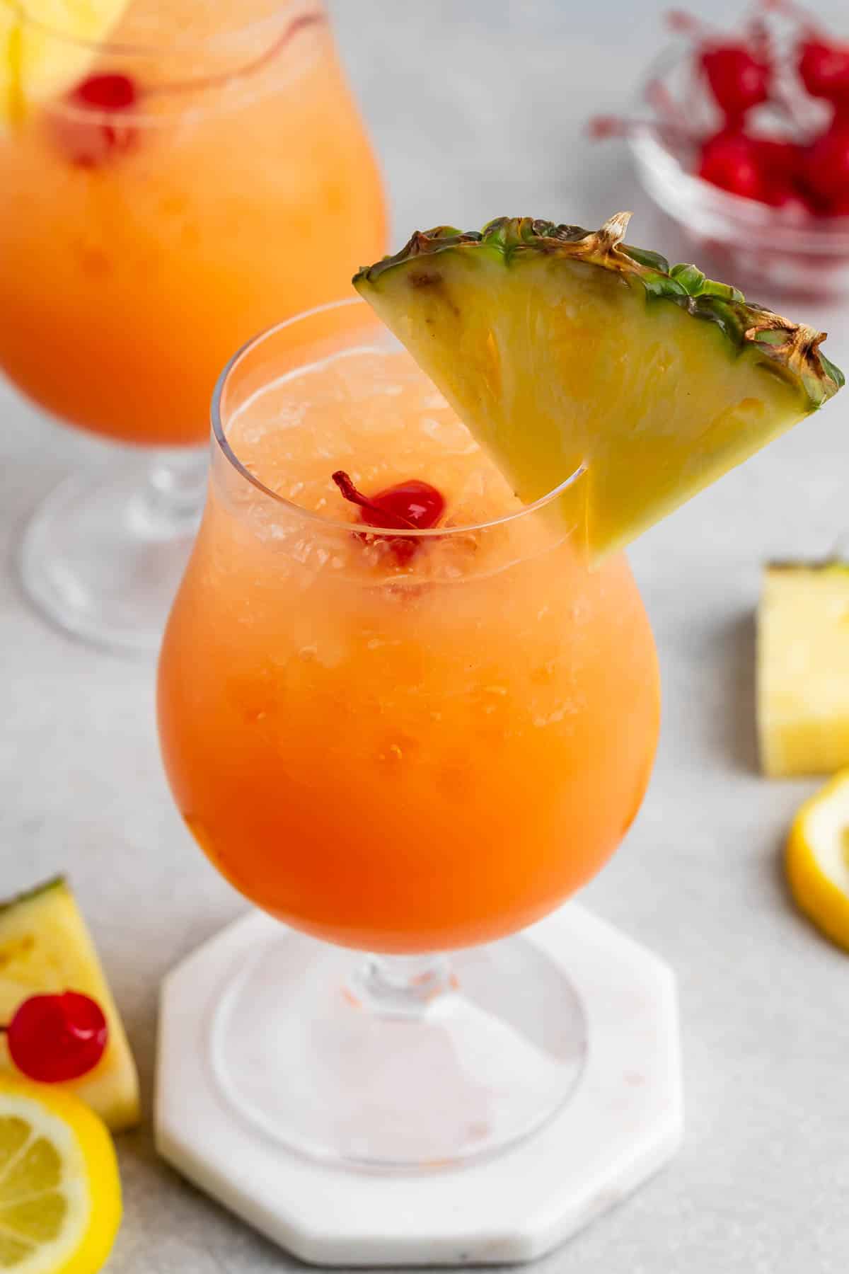 Two glasses of tropical cocktails with orange hue, garnished with pineapple slices and cherries. They are served in goblet-style glasses on a white coaster. Lemon slices and cherries are scattered on the gray surface.
