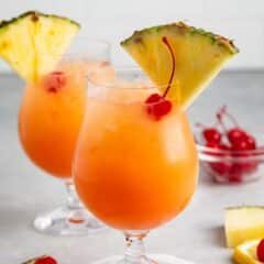 Two glasses of tropical cocktails with orange hue, garnished with pineapple slices and cherries. They are served in goblet-style glasses on a white coaster. Lemon slices and cherries are scattered on the gray surface.