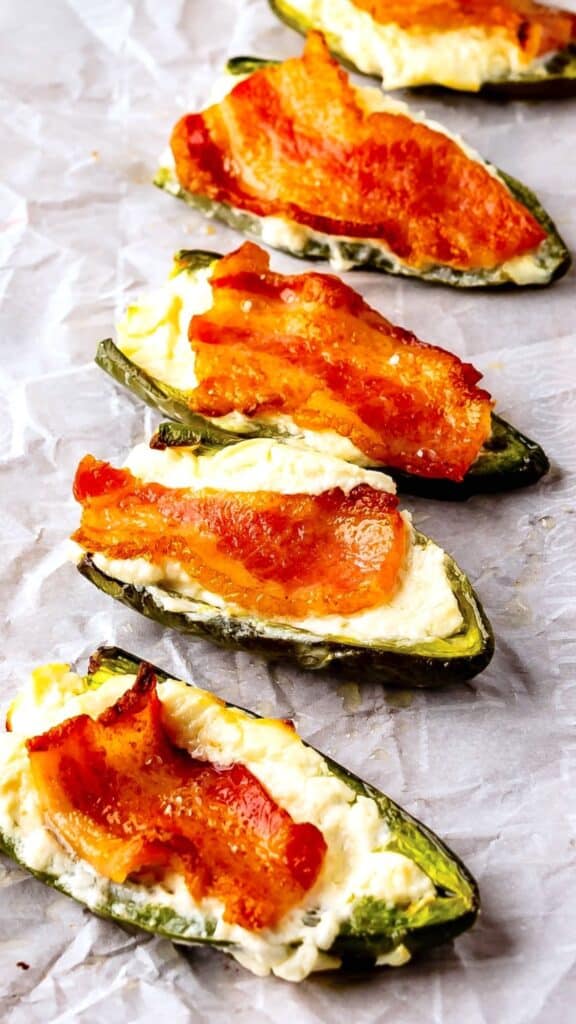jalapeños covered in cream cheese and bacon laid out on parchment paper.
