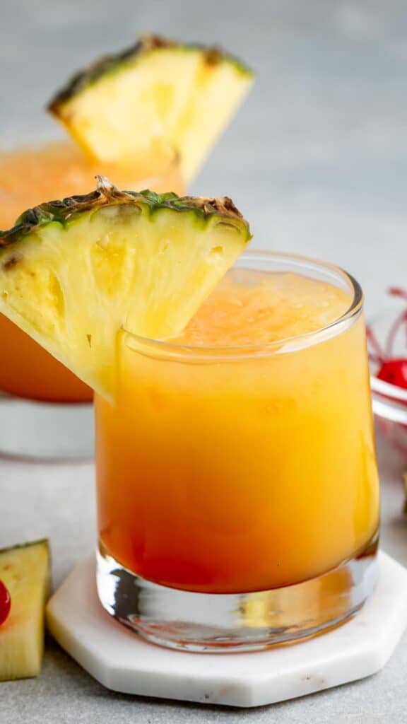 A glass of tropical orange cocktail garnished with a pineapple slice on the rim. The drink sits on a white coaster, with another similarly garnished drink and some cherries blurred in the background.