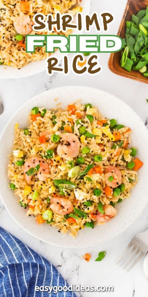 A plate of vibrant shrimp fried rice with peas, carrots, corn, and black sesame seeds. A fork rests on a striped napkin nearby. Fresh green onions are in the background. Text on the image reads Shrimp Fried Rice and easygoodideas.com.