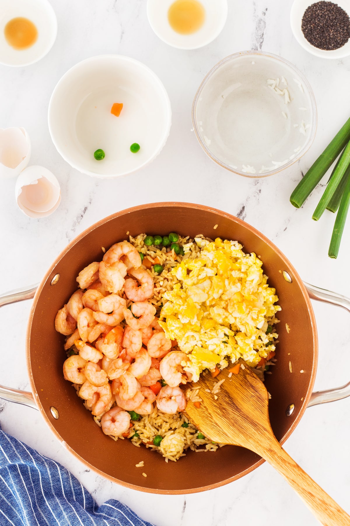 A copper pan on a marble countertop contains shrimp, scrambled eggs, and fried rice being stirred with a wooden spoon. Surrounding the pan are small bowls with ingredients and a blue striped cloth. Scallions are on the side.