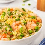 A bowl of shrimp fried rice with peas, carrots, scrambled eggs, and chopped green onions on top. The dish is placed on a marble surface with a blue-striped napkin, a wooden container, and stacked white plates in the background.