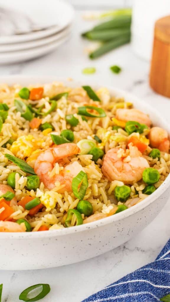 A close-up of a bowl filled with shrimp fried rice. The dish includes rice, shrimp, peas, carrots, and green onions, garnished with sliced green onions. A stack of plates and a blue napkin are in the background.
