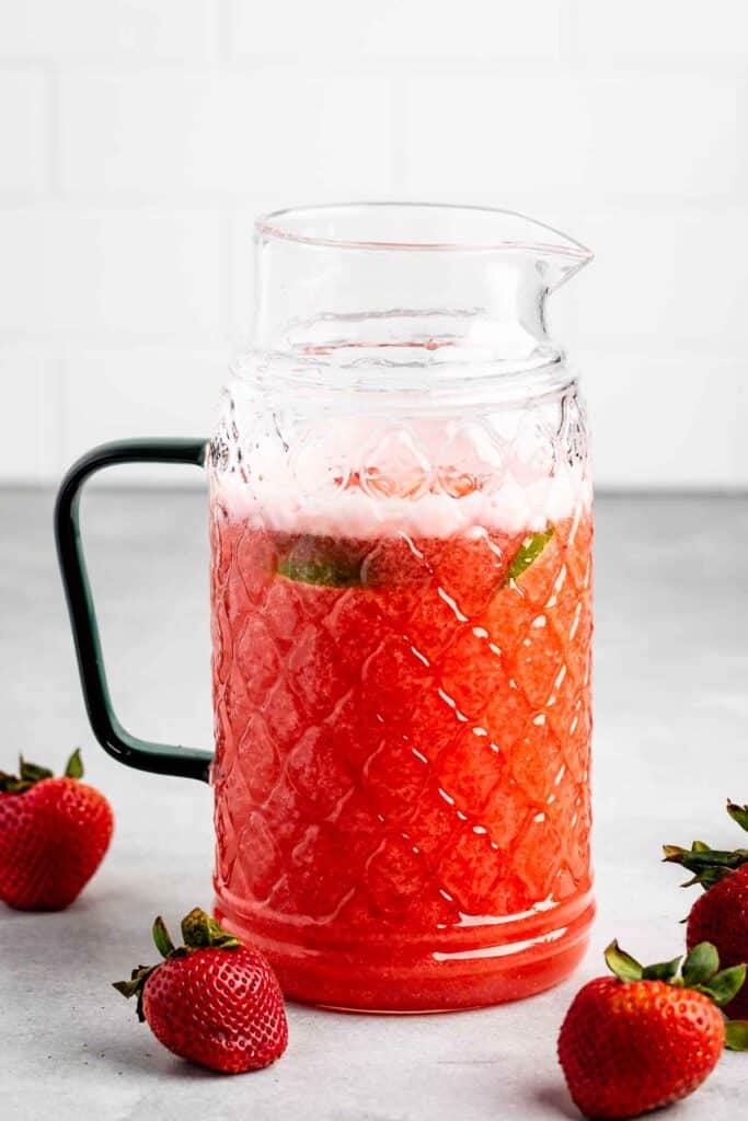 A glass pitcher filled with strawberry agua fresca, featuring a textured pattern and a handle. Fresh strawberries are placed around the pitcher.