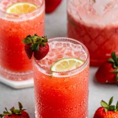 Two glasses of strawberry lemonade garnished with lime slices and fresh strawberries. The drinks are in textured glasses, and there are whole strawberries scattered around. In the background, theres a pitcher filled with the same beverage.