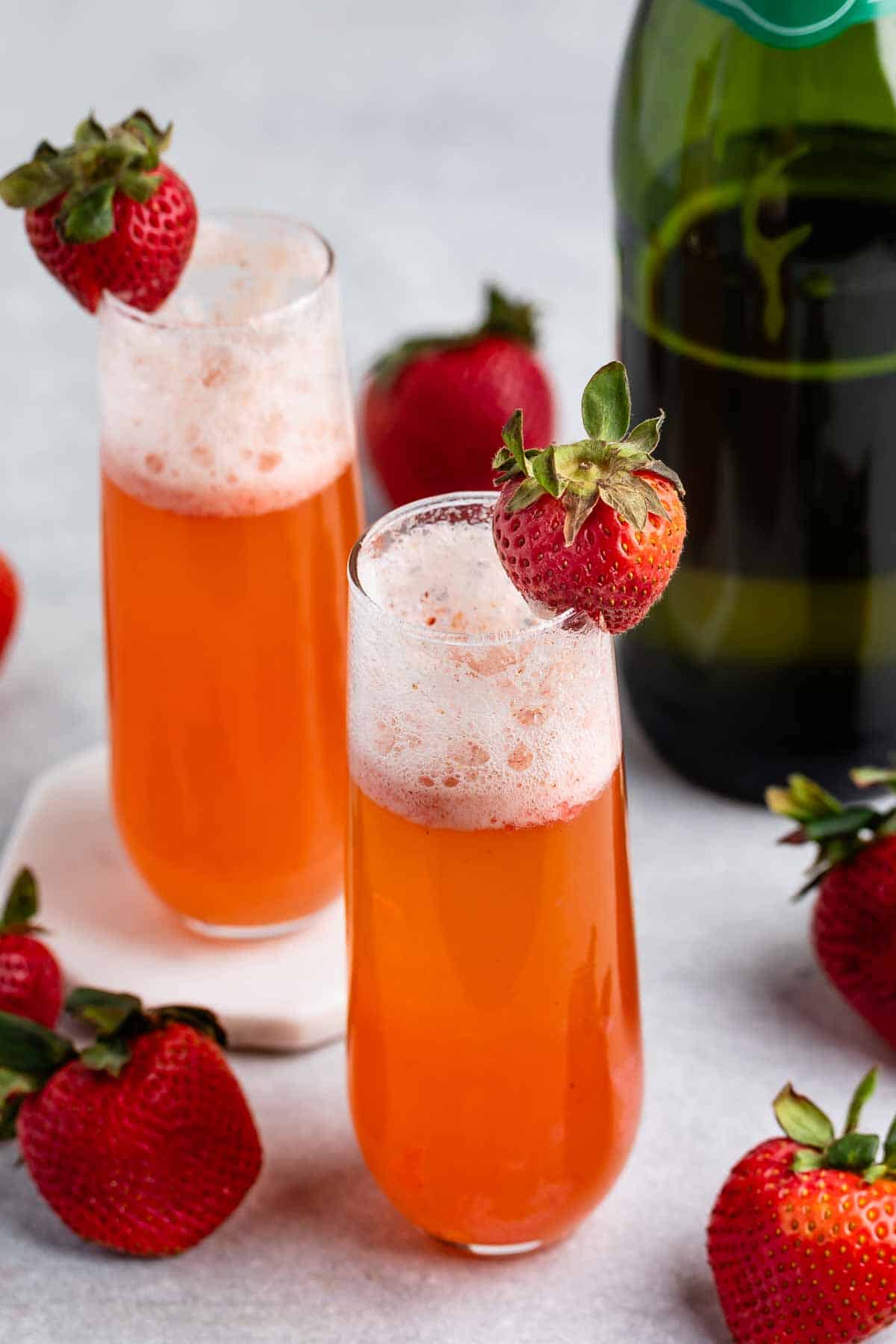 Two glasses of sparkling orange-colored beverage, each garnished with a fresh strawberry. A bottle stands in the background, surrounded by scattered strawberries on a light gray surface.