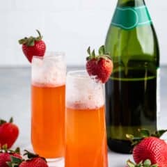 Two champagne flutes filled with a bubbly orange drink, each garnished with a ripe strawberry. A green bottle of champagne is in the background. Several strawberries are scattered around on a light surface.