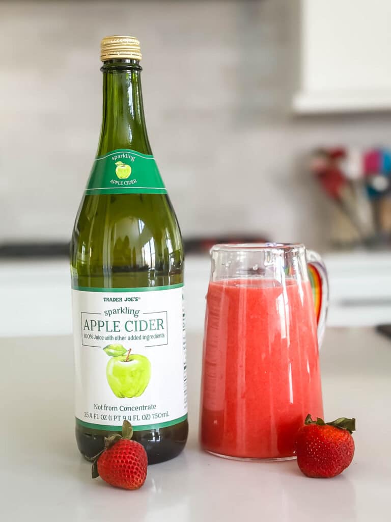 A bottle of sparkling apple cider is next to a glass pitcher filled with pink liquid. Two strawberries are placed beside the pitcher on a white countertop in a kitchen setting.