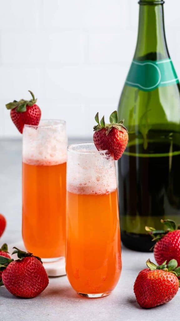 Two tall glasses filled with a fizzy orange beverage, each garnished with a strawberry on the rim. A green bottle is in the background, with strawberries scattered around on a light surface.