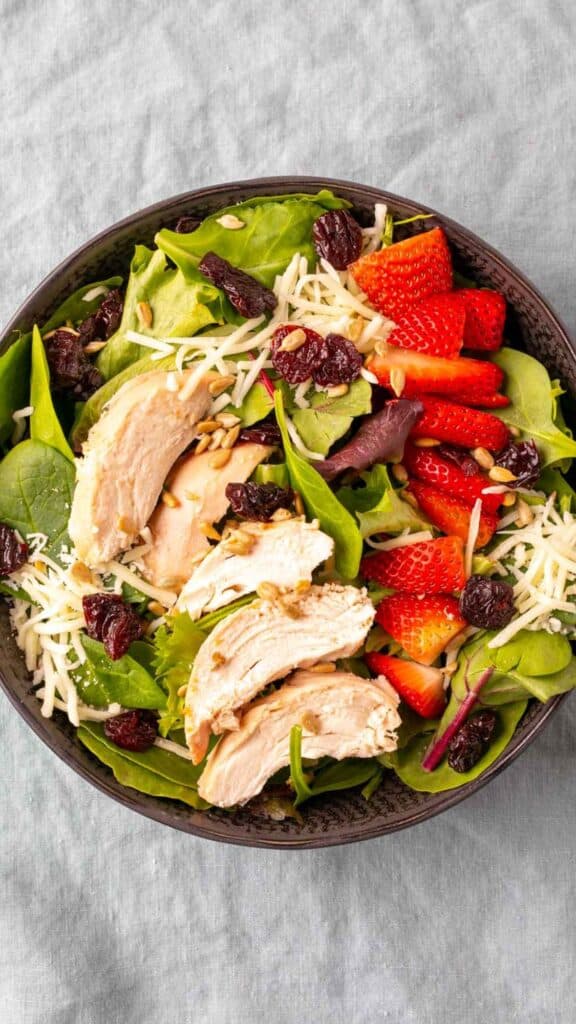 A bowl of salad with sliced grilled chicken, fresh strawberries, shredded cheese, mixed greens, dried cranberries, and sunflower seeds, set on a light gray tablecloth.