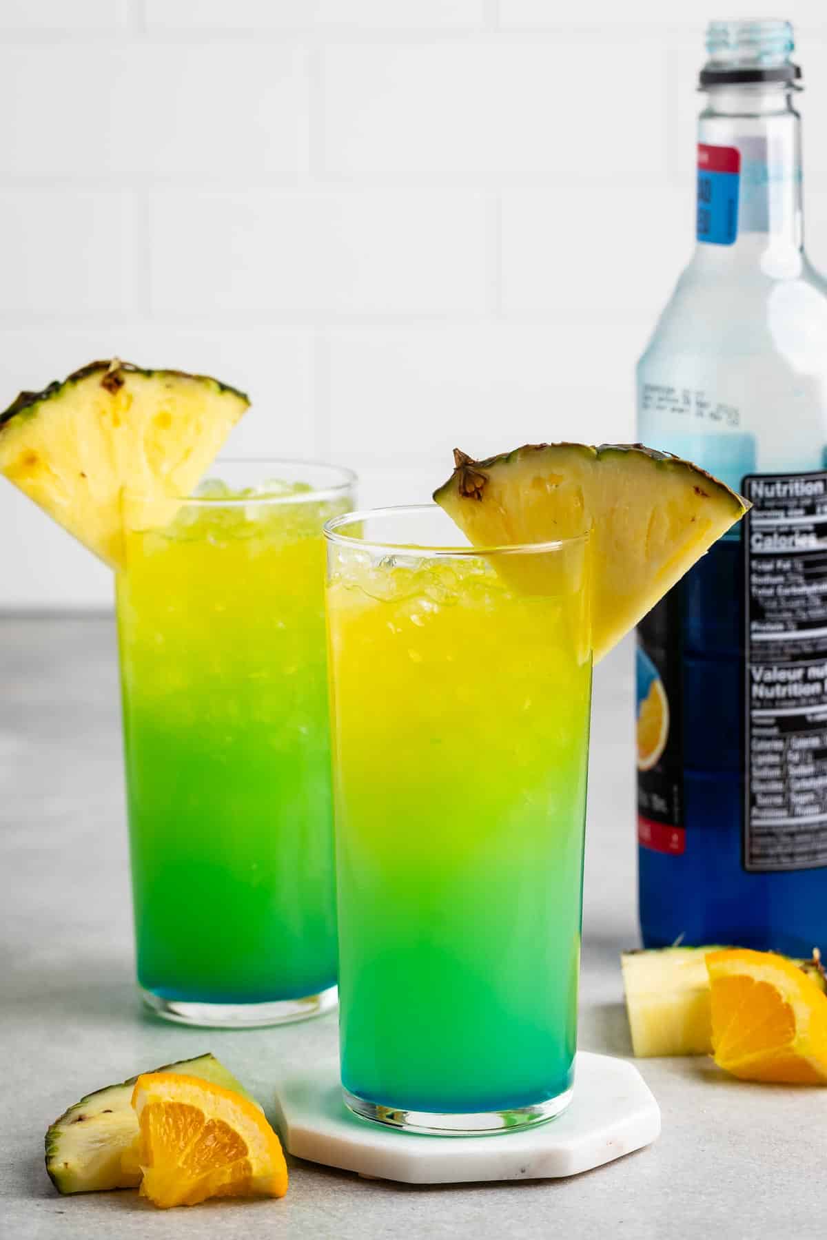 Two tall glasses filled with a vibrant blue and green layered drink, garnished with pineapple wedges. A bottle of blue liquid is in the background. Orange slices and pineapple pieces are scattered around on a light gray surface.