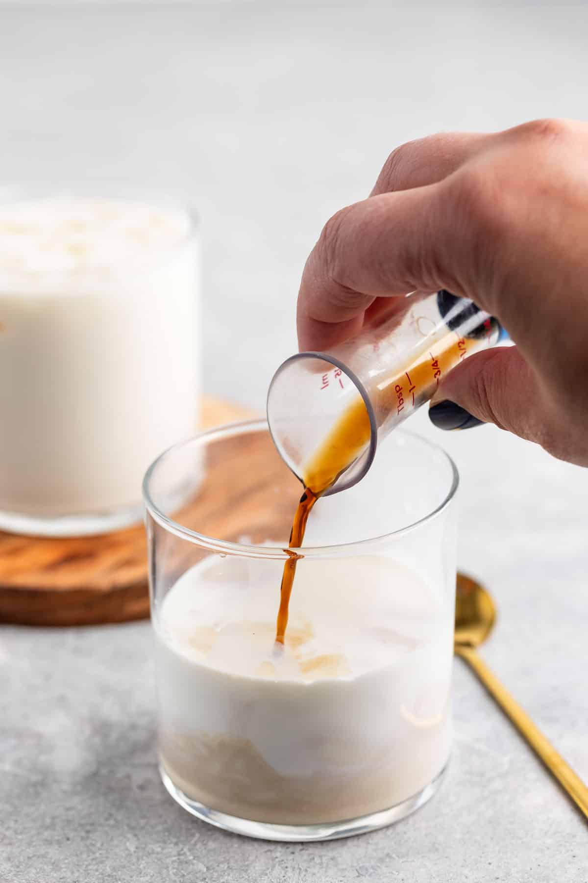 A hand pours espresso from a small measuring glass into a clear glass filled with milk. A frothy swirl forms in the milk. Another similar drink and a gold spoon sit on a wooden coaster and a gray surface in the background.