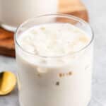 A glass filled with iced milk tea, featuring small ice cubes. A gold spoon lies nearby on a gray surface. A wooden coaster with another glass is partially visible in the background.