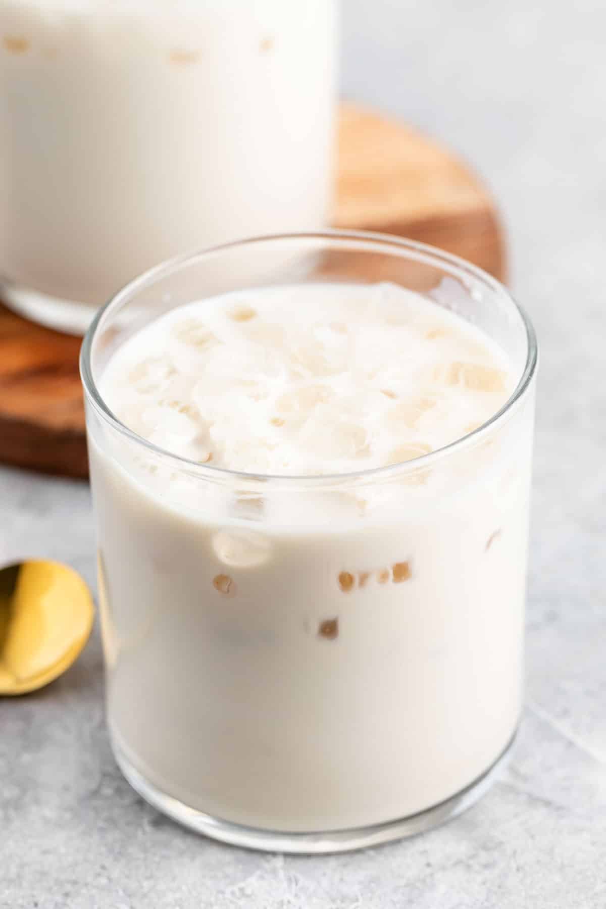 A glass filled with iced milk tea, featuring small ice cubes. A gold spoon lies nearby on a gray surface. A wooden coaster with another glass is partially visible in the background.