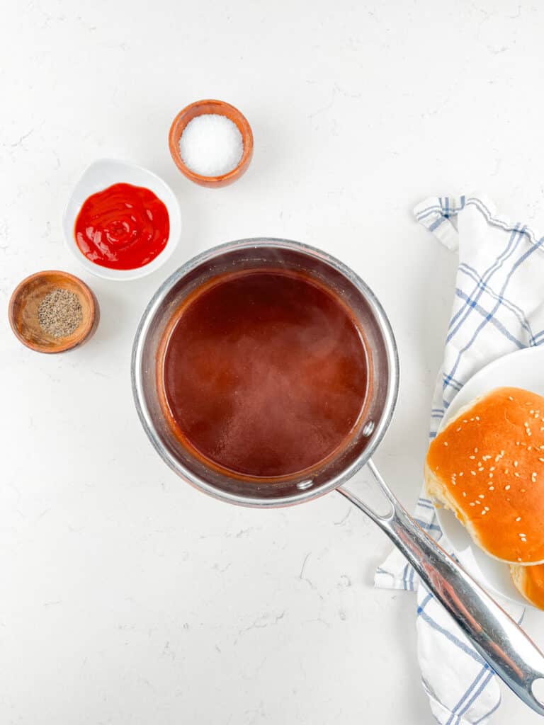 A saucepan filled with red sauce sits on a white countertop next to small bowls of salt, ketchup, and pepper. A sesame seed bun and a folded checkered cloth are nearby.
