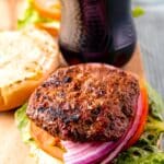 A juicy grilled burger patty rests on an open bun with lettuce, tomato, and sliced red onion. In the background, theres a bottle of Coca-Cola with a visible label. The setup is on a wooden surface, suggesting a casual meal setting.