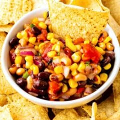 A white bowl filled with colorful corn, black bean, tomato, and onion salsa, surrounded by yellow tortilla chips, with one chip dipped into the salsa.