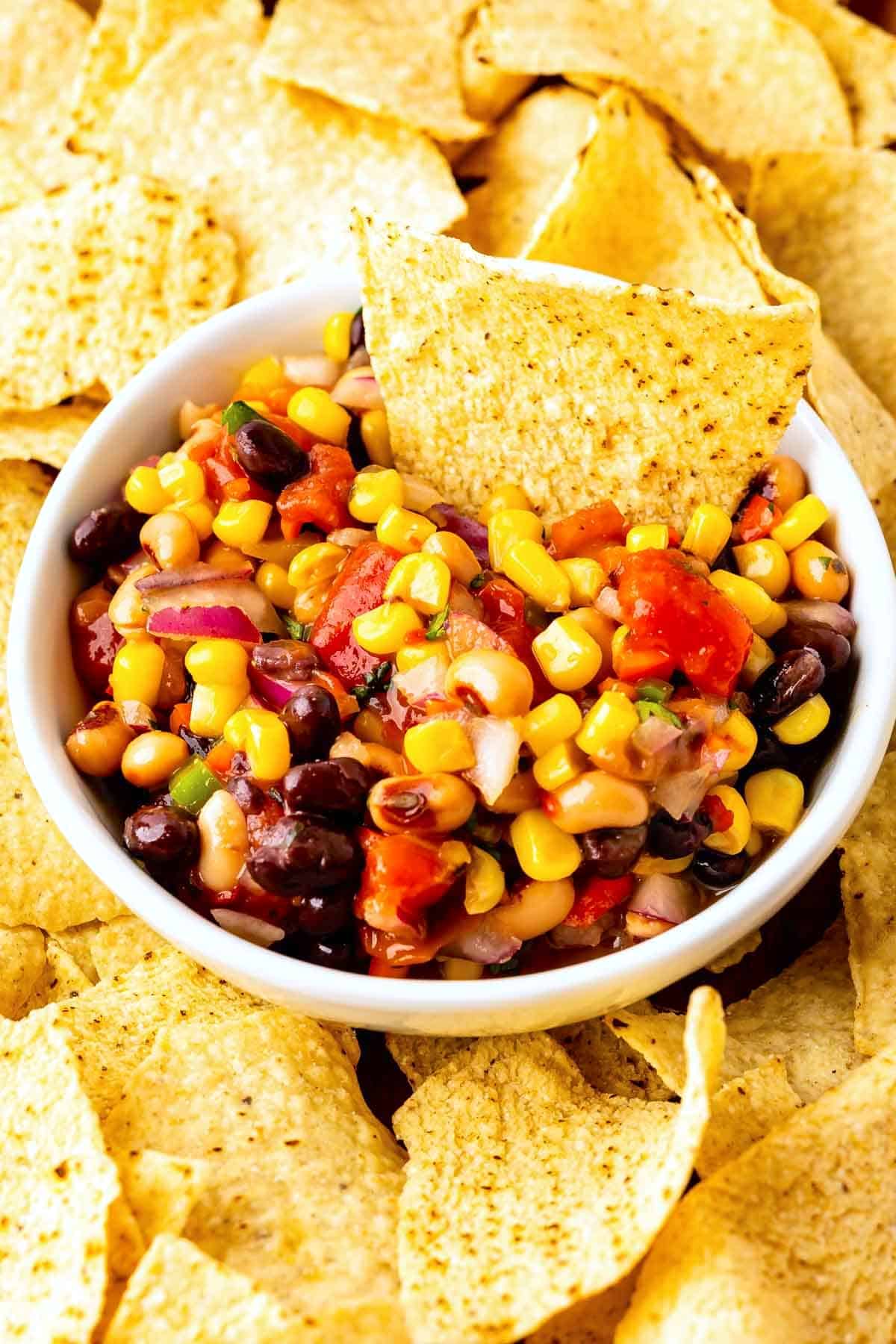 A white bowl filled with colorful corn, black bean, tomato, and onion salsa, surrounded by yellow tortilla chips, with one chip dipped into the salsa.