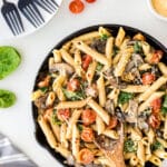 A skillet filled with creamy penne pasta, mushrooms, spinach, and cherry tomatoes sits on a white surface with a wooden spoon, surrounded by plates, forks, spinach leaves, grated cheese, chili flakes, and whole cherry tomatoes.