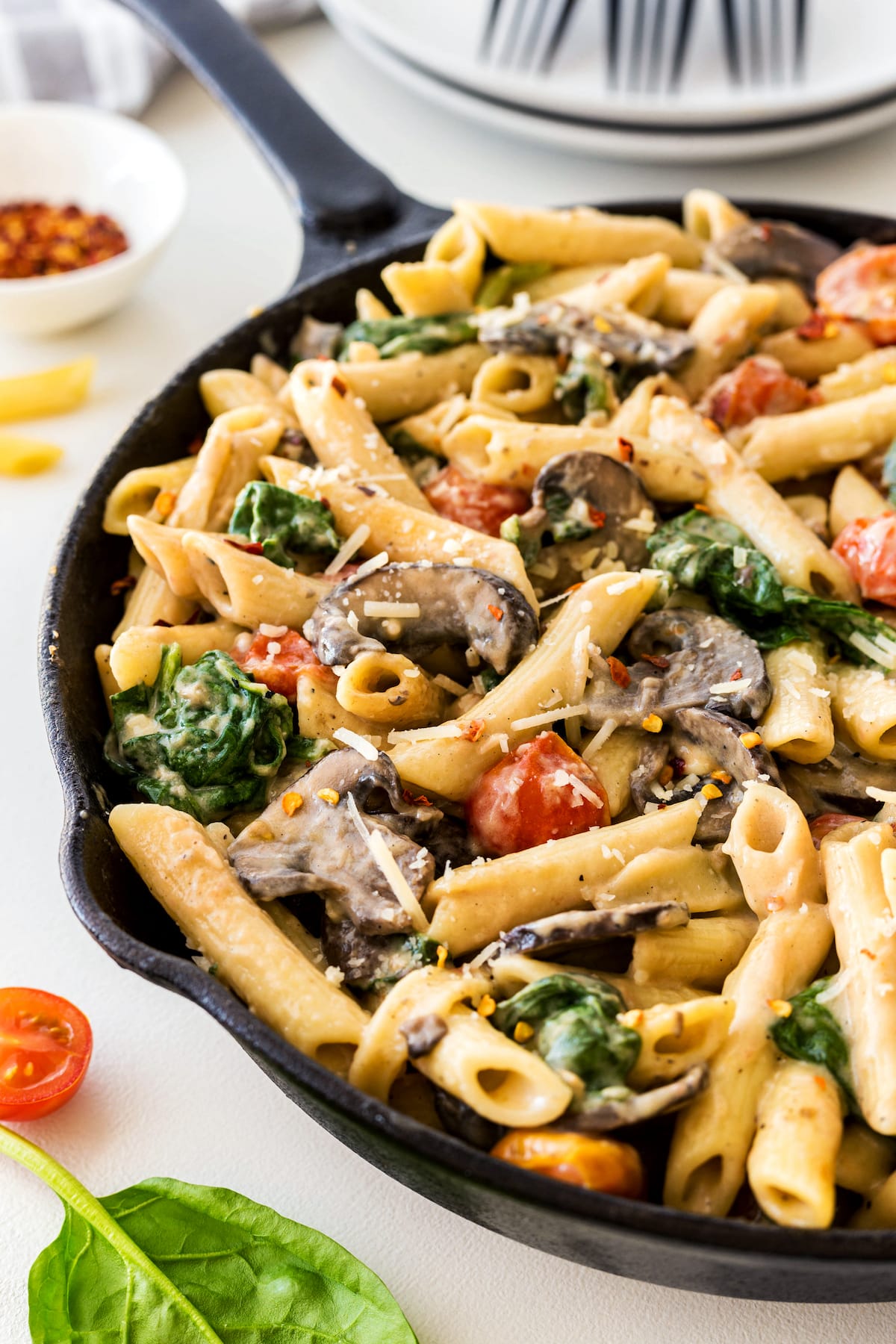 A black skillet filled with creamy penne pasta, mushrooms, cherry tomatoes, spinach, and topped with grated parmesan cheese sits on a white table with a plate and utensils in the background.