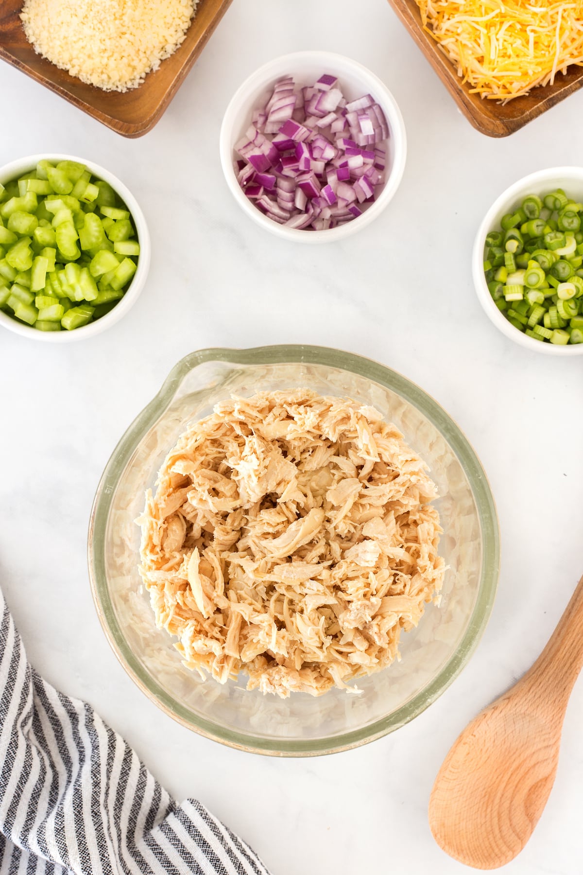 A glass bowl of shredded chicken is centered on a countertop. Surrounding it are small bowls of chopped green bell pepper, sliced green onions, diced red onion, shredded cheese, and breadcrumbs. A wooden spoon and striped cloth are nearby.
