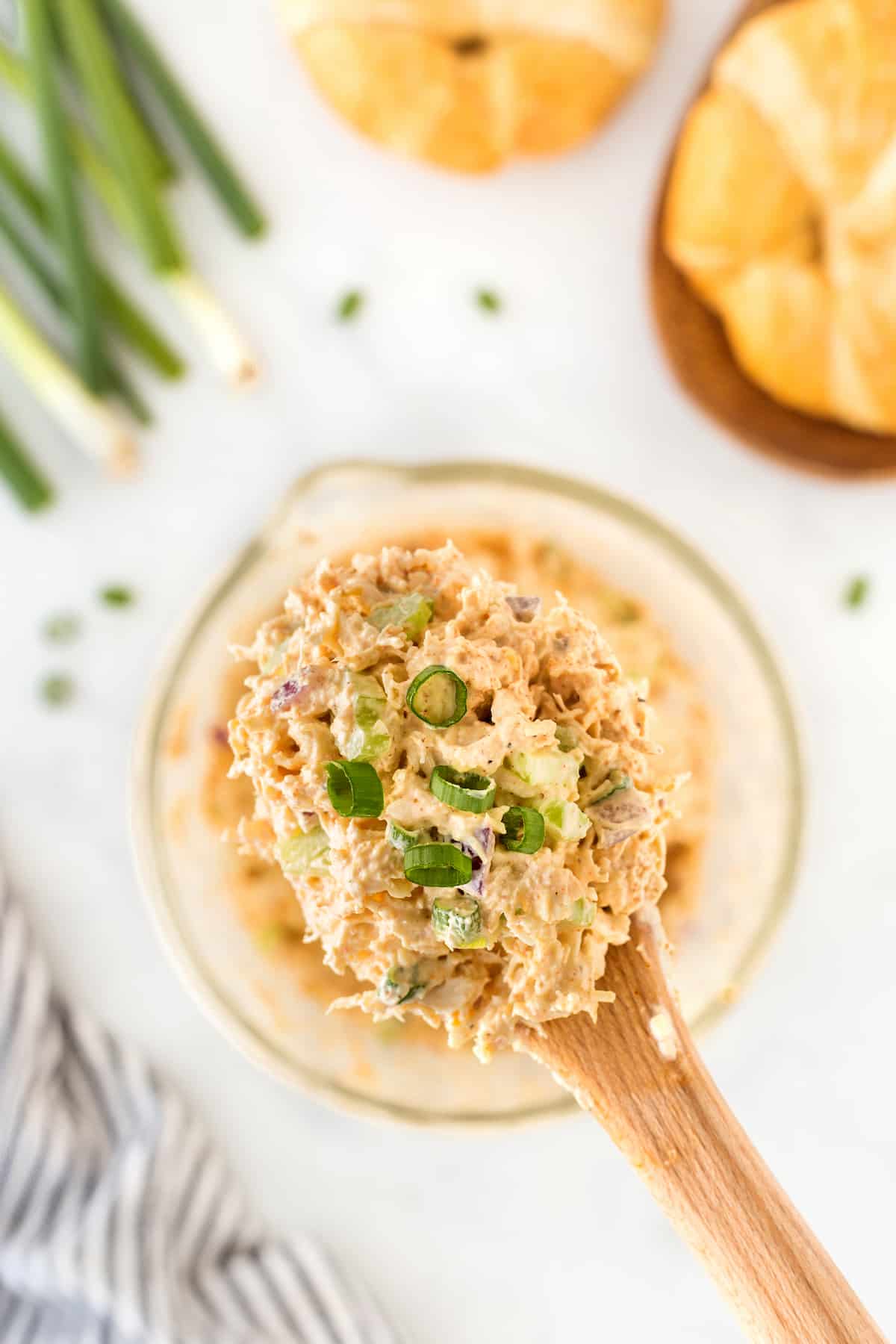 A wooden spoon holds a scoop of chicken salad garnished with sliced green onions over a bowl. Croissants and green onions are in the background. A striped cloth is partially visible on the side.