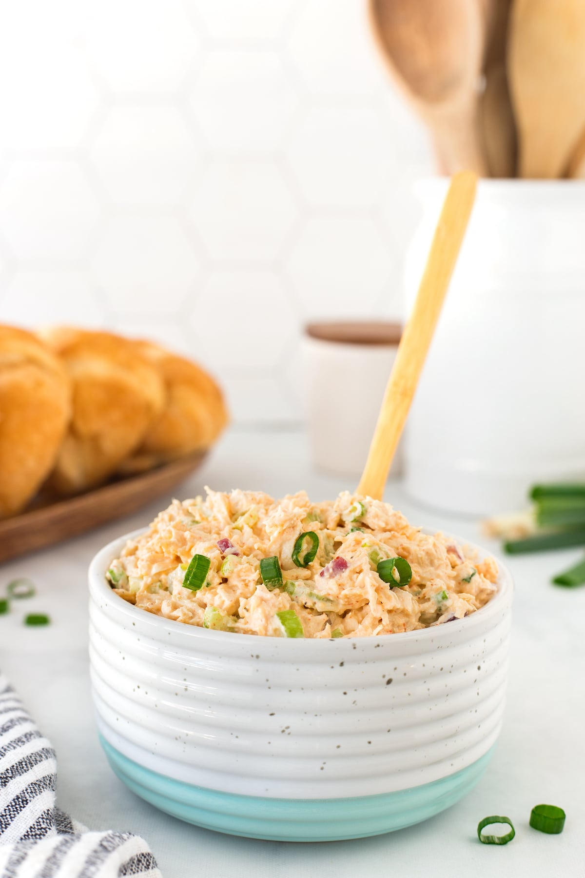 A ceramic bowl with a speckled design, filled with creamy pimento cheese and garnished with chopped green onions. A wooden spoon is inserted into the mixture. In the background, fresh bread rolls and kitchen utensils are visible.