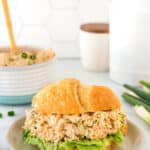 A croissant sandwich filled with chicken salad and lettuce is placed on a plate. In the background, a bowl of chicken salad and kitchen utensils are visible. The setting has a light, airy feel with a hexagonal-tiled wall.