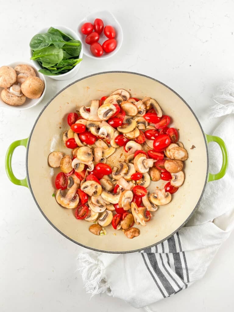 A green-handled pot filled with sliced mushrooms and halved cherry tomatoes sits on a white surface. Nearby are small bowls of fresh spinach, whole mushrooms, and cherry tomatoes. A striped towel is beside the pot.