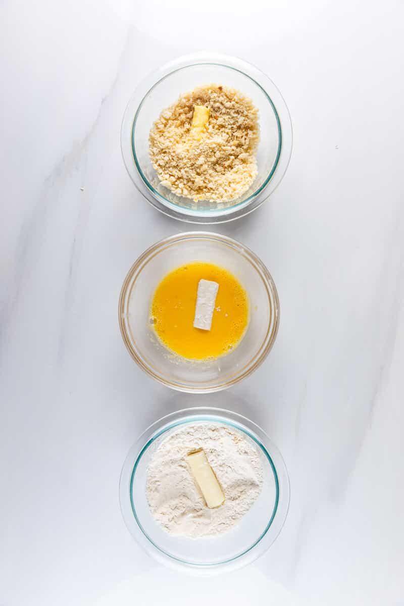 Three glass bowls on a marble surface contain flour with butter, beaten eggs, and crushed mixture with butter, respectively. These ingredients are typically used for breading or coating preparations.