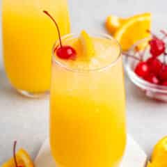 Two glasses of orange juice with ice, each garnished with a cherry and an orange slice, are set on a light surface. A small bowl of cherries and orange wedges are in the background. A hexagonal coaster holds one glass in the foreground.