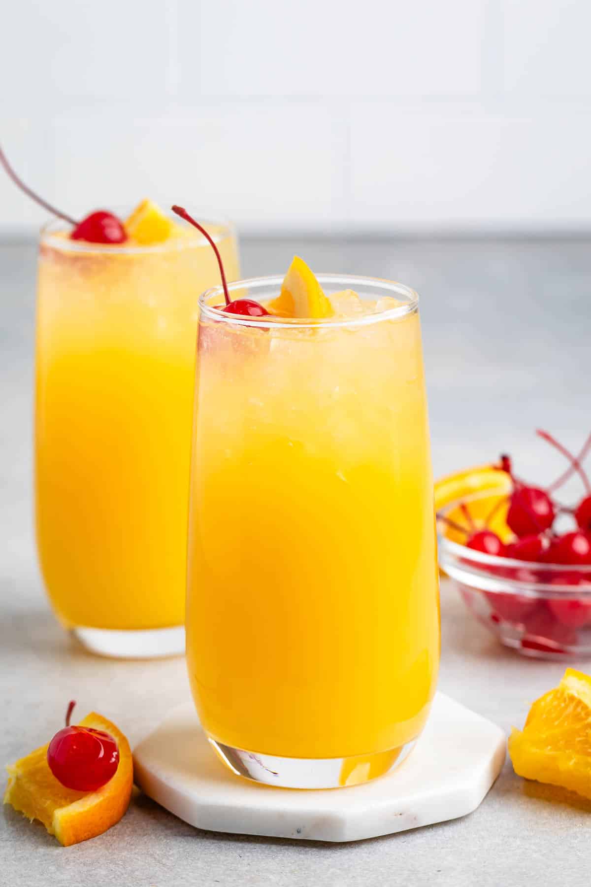 Two glasses of vibrant orange juice garnished with cherries and orange slices sit on a light countertop. In the background, a small bowl holds more cherries and orange wedges.