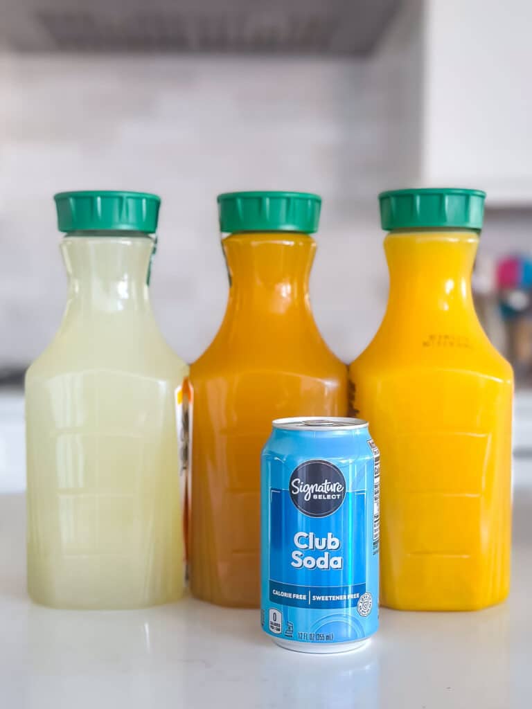Three large bottles of juice with green caps and a can of club soda. The bottles contain different colored juices: light yellow, bright orange, and medium orange. The can is blue and labeled Signature Select Club Soda. They are on a kitchen counter.