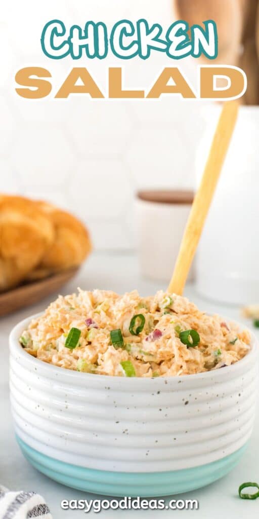 A bowl of creamy chicken salad garnished with chopped green onions, with a wooden spoon in it. Fresh bread and a white cup are in the blurred background. The words Chicken Salad are displayed at the top.