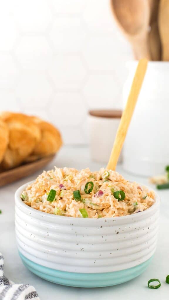 Bowl of creamy shredded chicken salad garnished with chopped green onions. A wooden spoon is inside the bowl. In the background, blurred rolls on a plate and kitchen utensils are visible. The scene is set on a light counter.