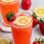 Two tall glasses of refreshing strawberry lemonade, each garnished with a lemon slice and a fresh strawberry. Surrounding the glasses are whole strawberries and lemon slices on a light background.