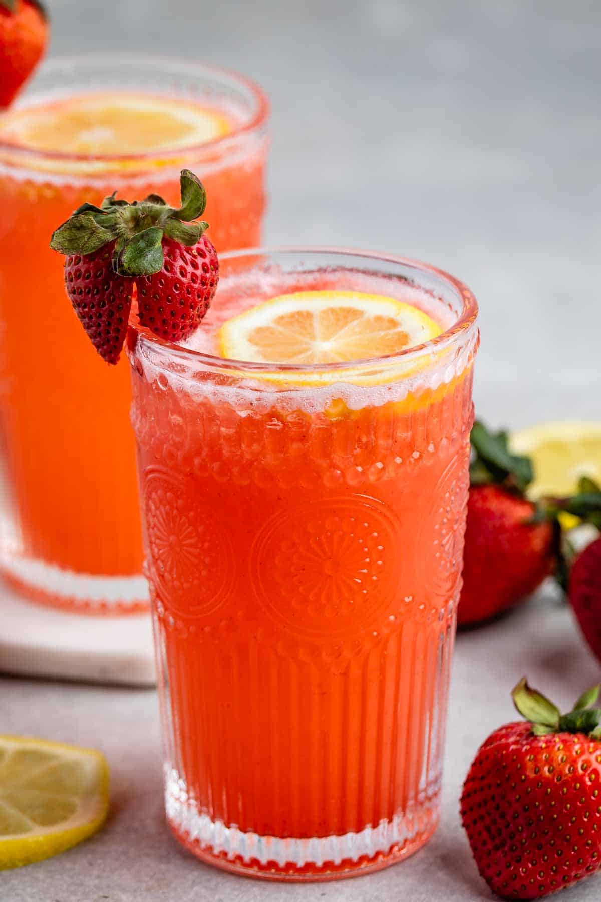 Two textured glasses filled with vibrant orange-red liquid, topped with lemon slices. Each glass has a fresh strawberry on the rim. Strawberries and lemon slices are scattered around on a light background.