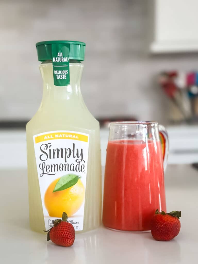 A bottle of Simply Lemonade is next to a glass pitcher of bright red strawberry lemonade. Two strawberries are placed in front, on a white countertop, with a blurred kitchen background.