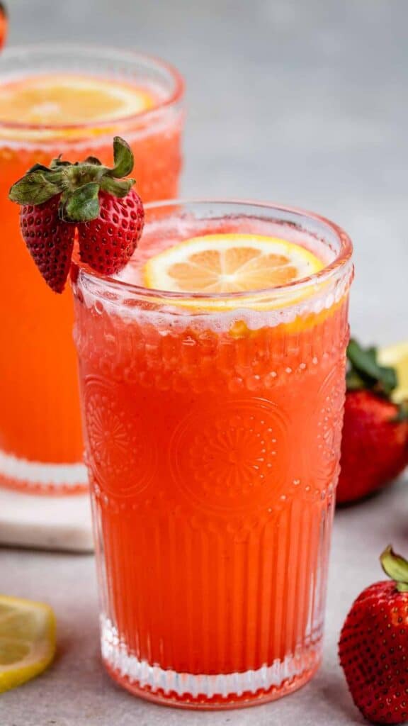 A close-up of two glasses of bright red strawberry lemonade, each garnished with a slice of lemon and a fresh strawberry on the rim. More strawberries and lemon slices are scattered around the glasses on a light surface.