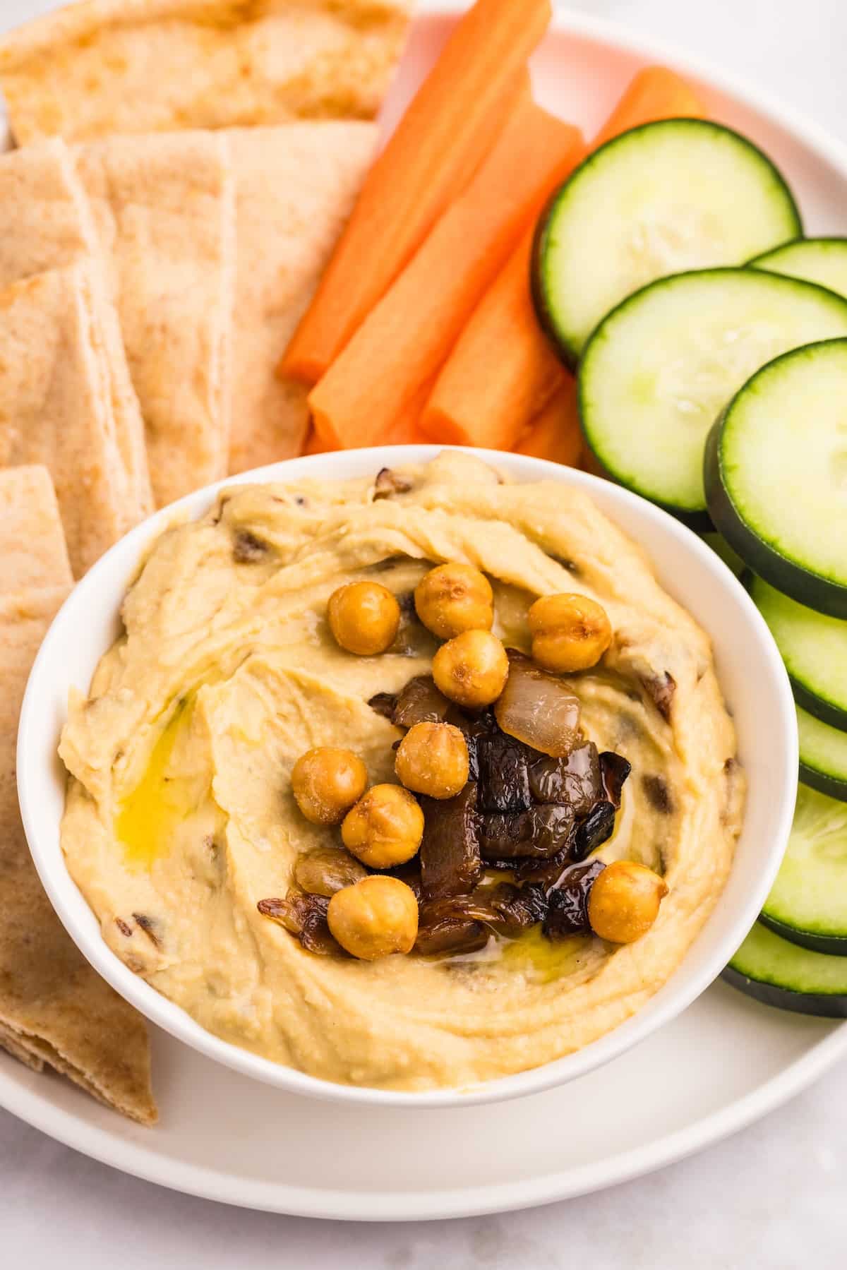A bowl of hummus topped with chickpeas and caramelized onions is surrounded by pita bread slices, carrot sticks, and cucumber slices on a white plate.